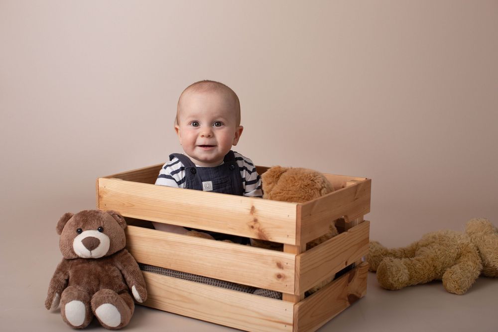 Adorable baby sitting inside wooden crate surrounded by teddy bears in professional studio photo.
