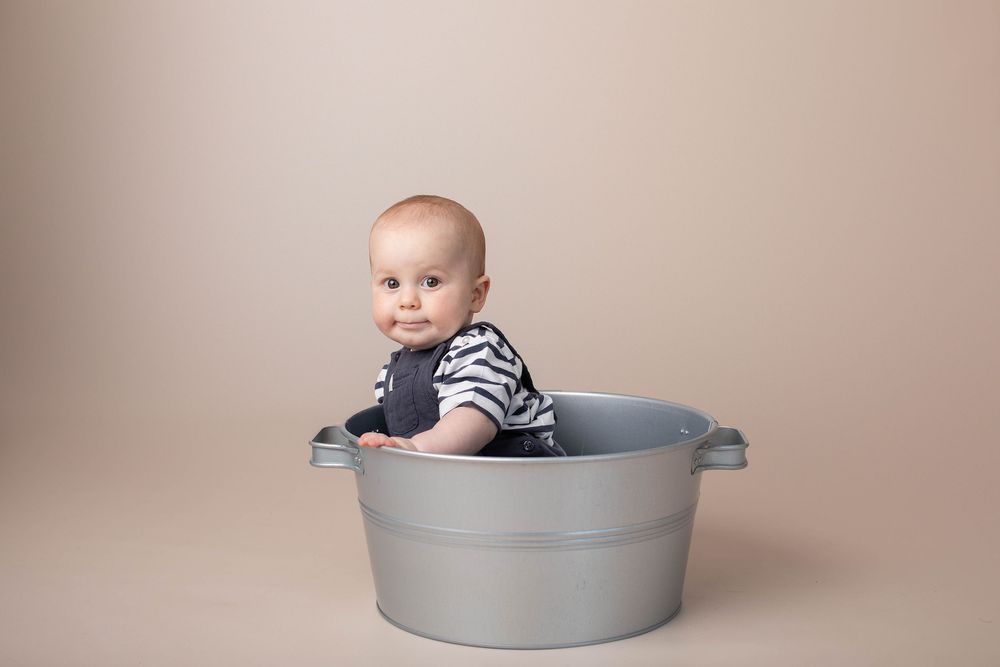 Cute baby sitting in silver metal tub during fun studio portrait session on beige background.