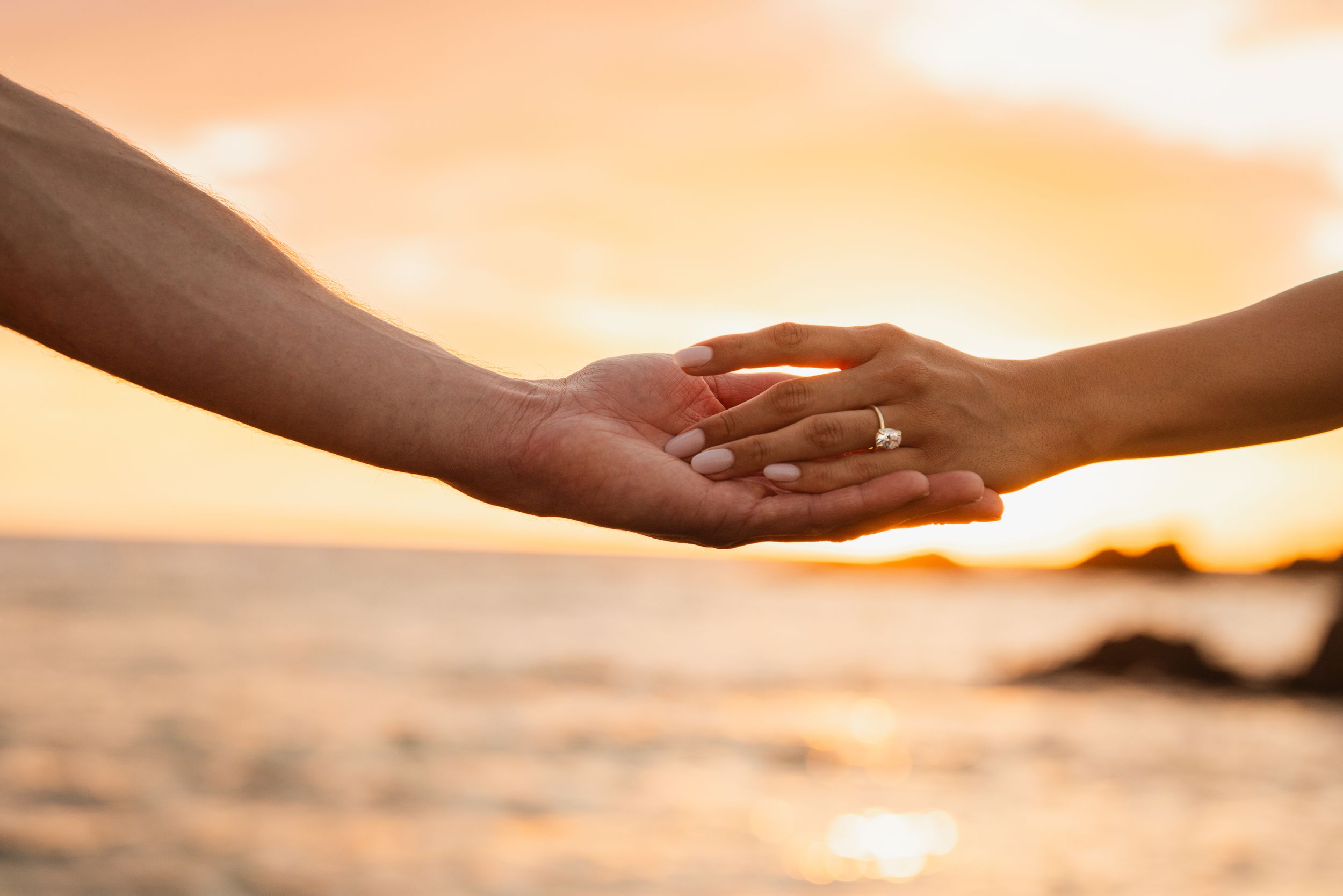Couple holding hands with engagement ring at sunset on beach.