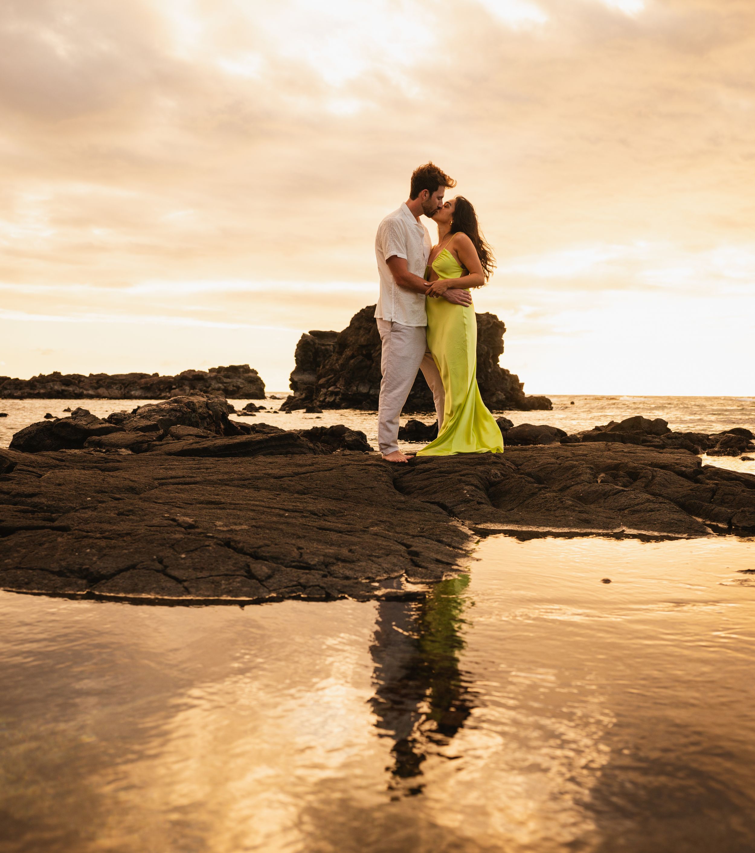 Couple embracing on rocky beach at sunset, woman in green gown, man in white, with ocean reflection below.