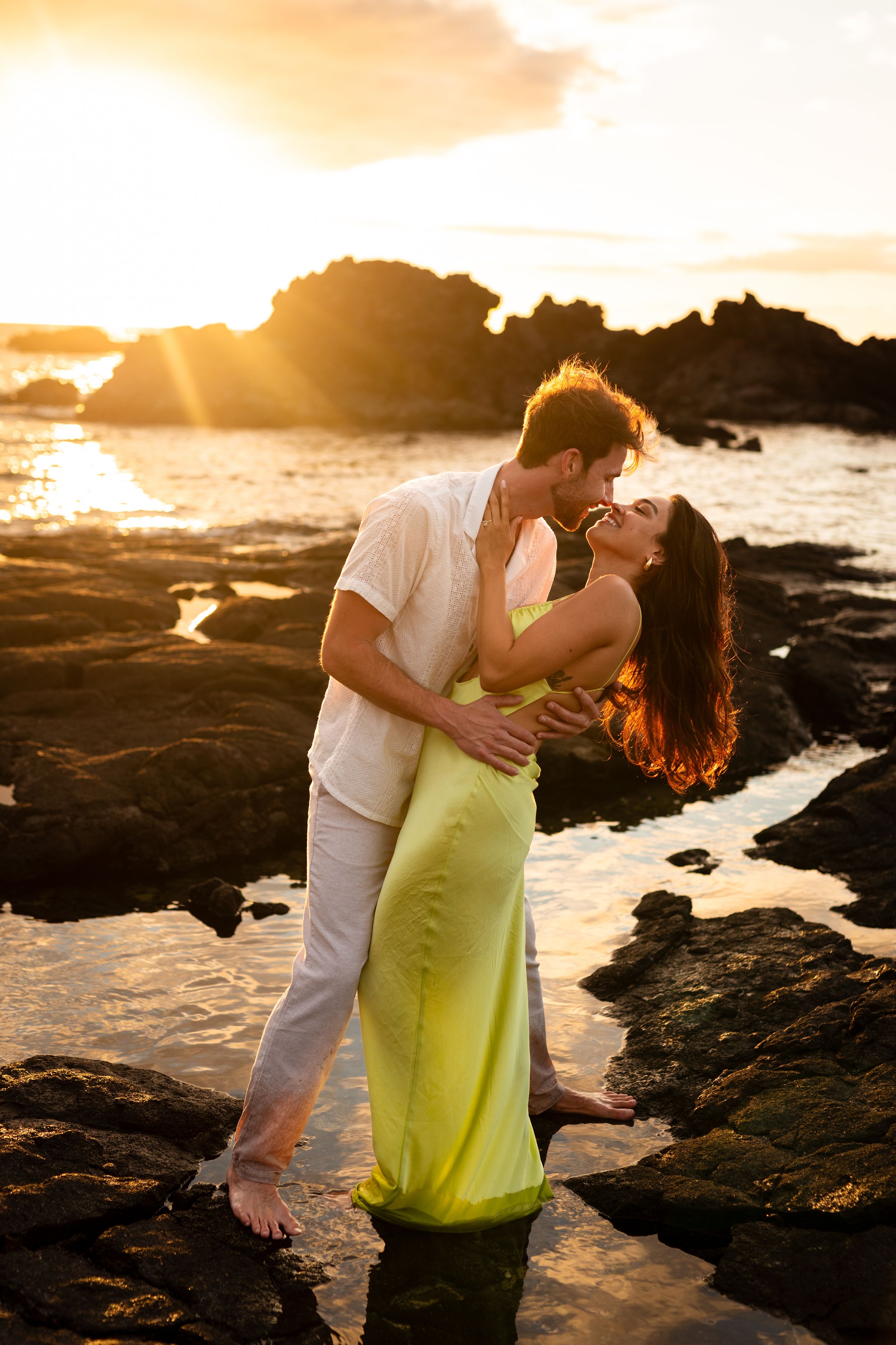 Couple embracing romantically on rocky beach at golden sunset, woman in yellow dress, man in white shirt.