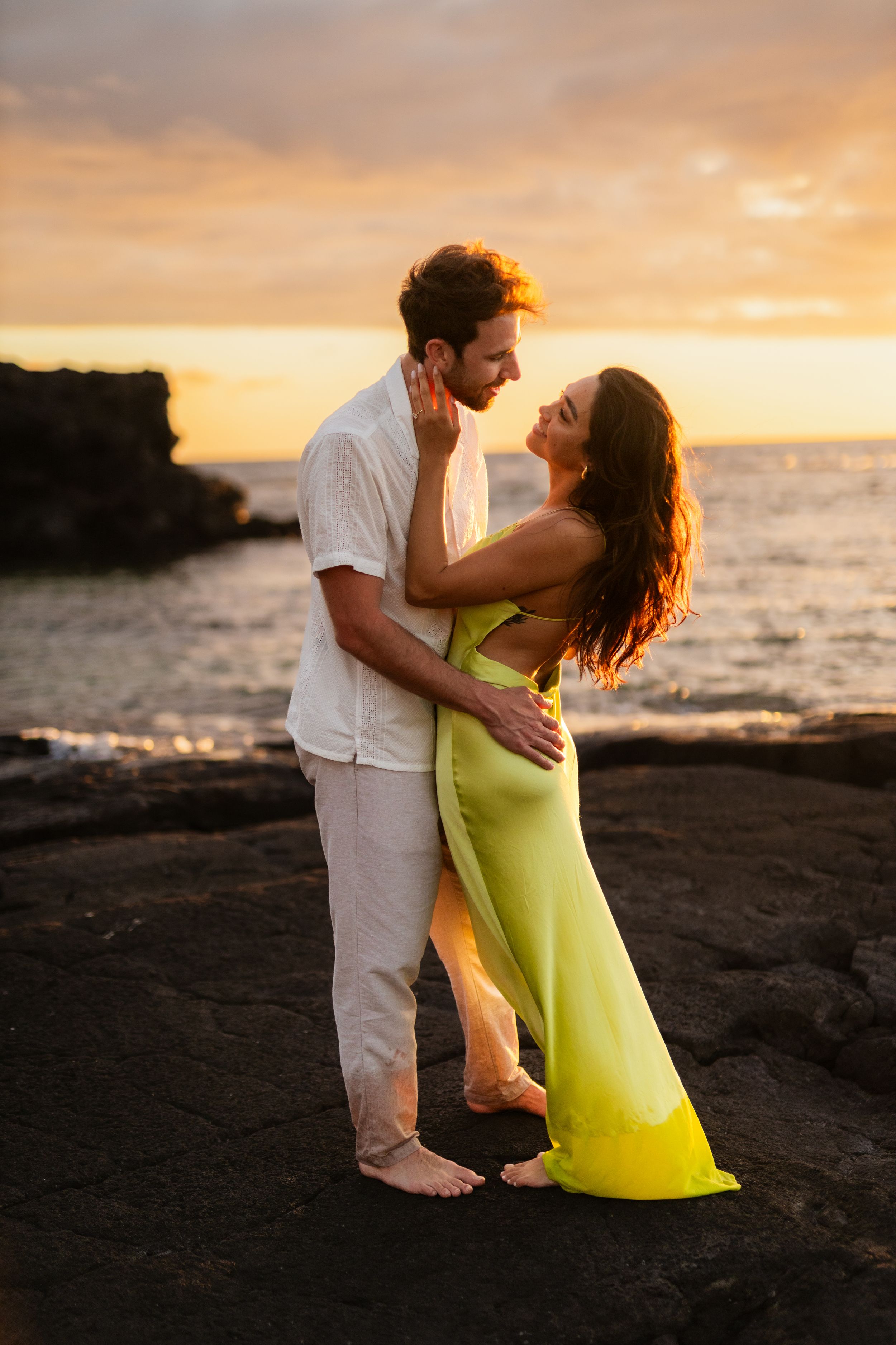 Couple embracing at sunset on rocky Hawaiian beach, woman in green dress, golden sky backdrop.