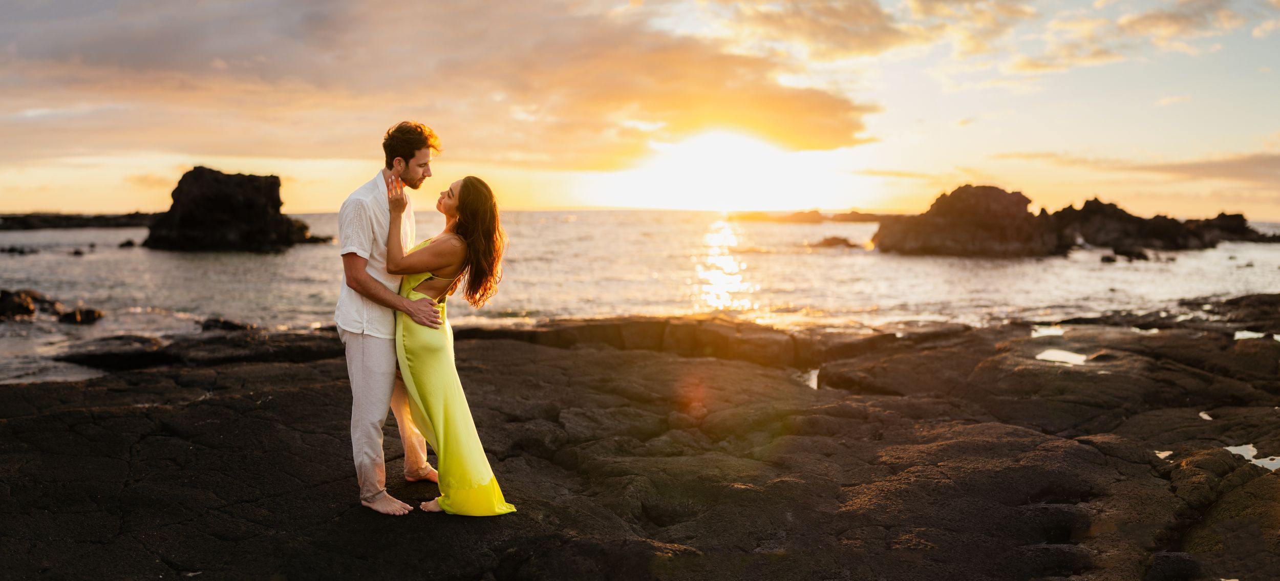 Couple embracing at sunset on rocky Hawaii beach, woman in yellow dress, golden light reflecting on ocean waves.