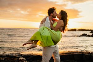 Couple sharing romantic kiss at sunset on rocky beach, man lifting woman in green dress with golden sky backdrop.