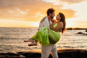 Romantic couple embracing at sunset on rocky beach, man carrying woman in green dress with ocean backdrop.