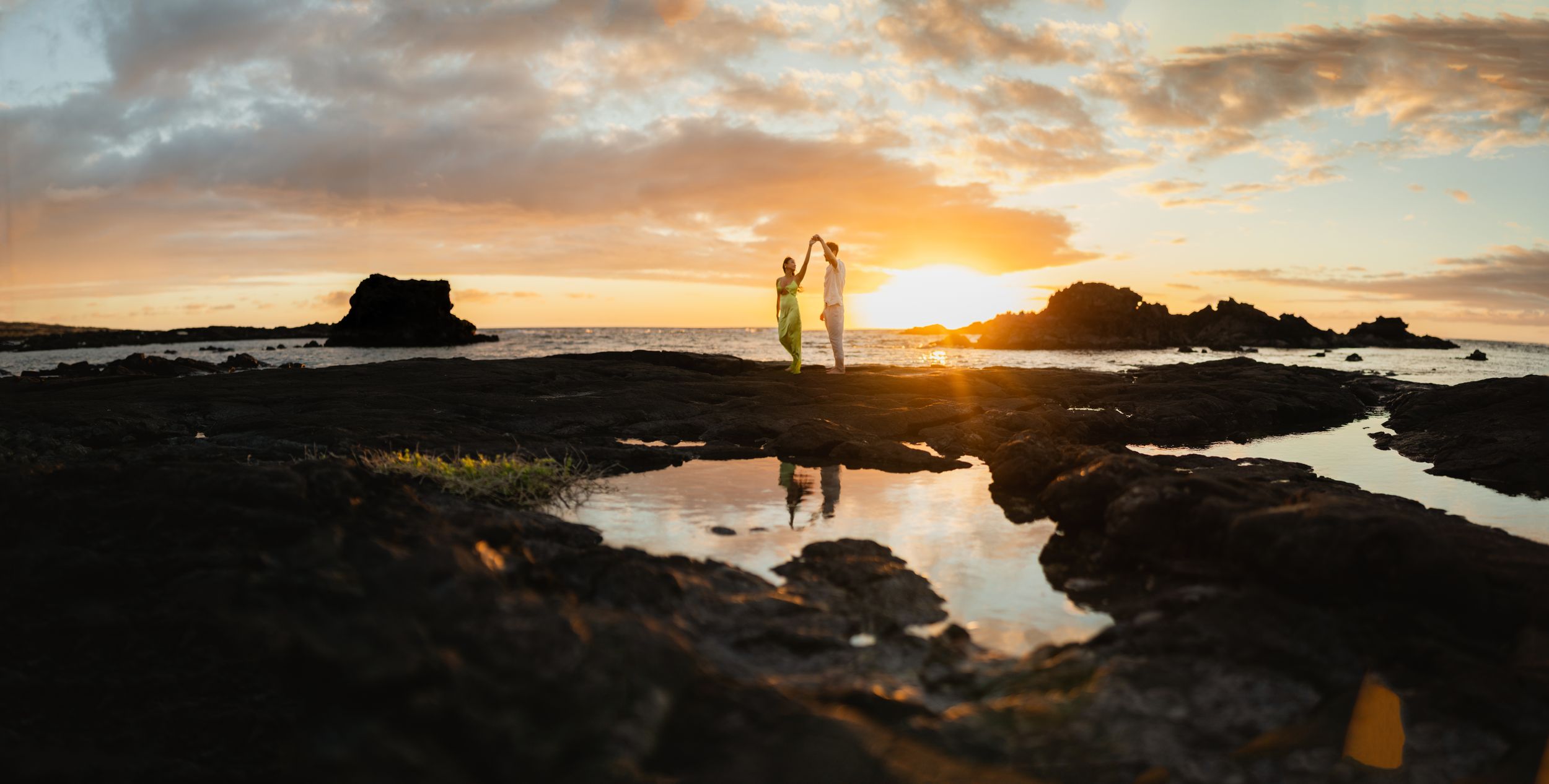 Couple dancing on rocky shore at sunset with golden reflections in tide pools and dramatic sky backdrop.