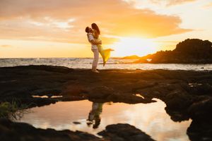 Couple embracing at sunset on rocky Hawaii beach, golden sky reflecting in tide pools below.