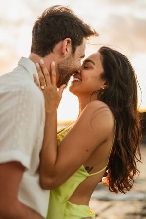 Romantic couple sharing a tender kiss at sunset on the beach, woman in yellow bikini top smiling.