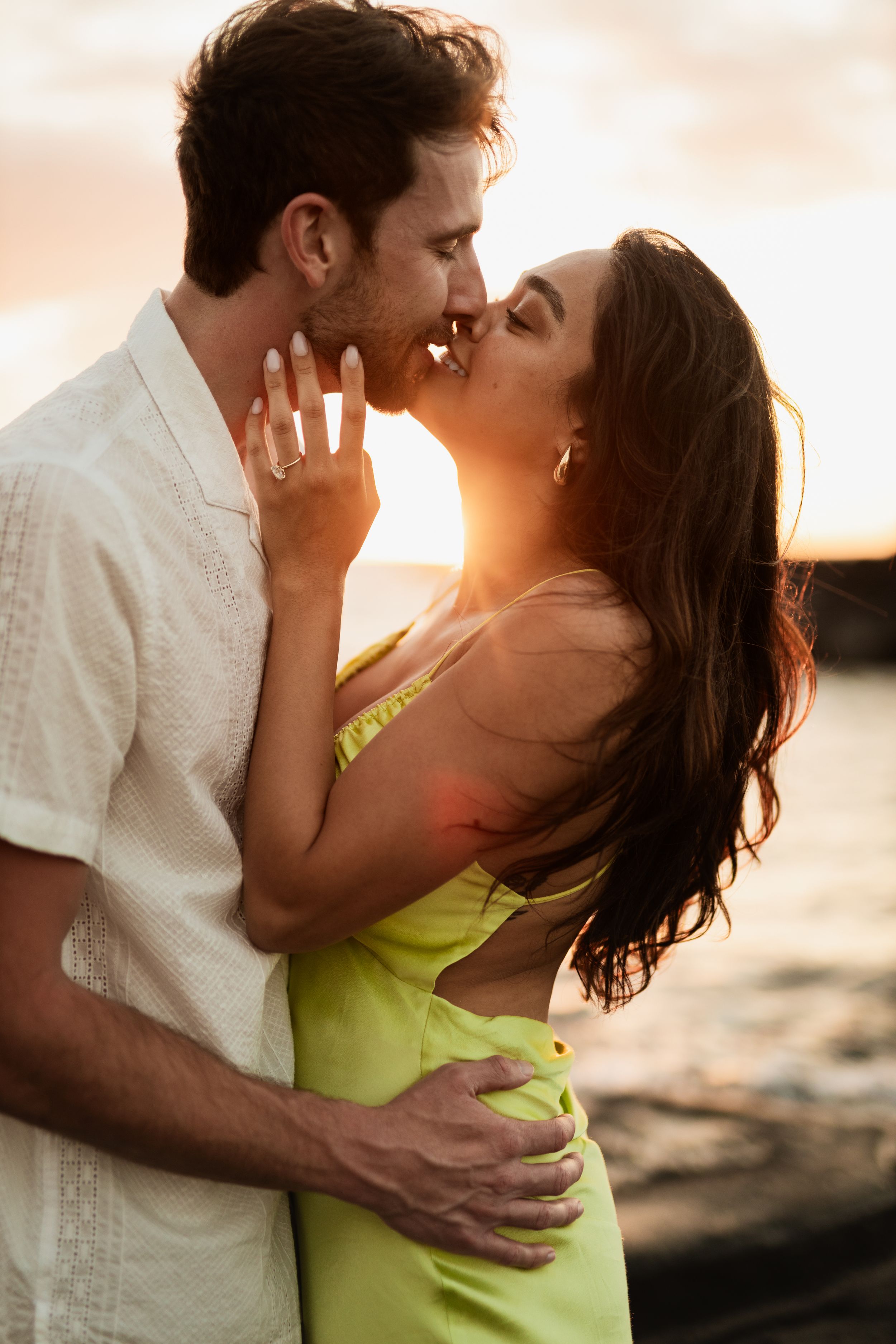 Couple sharing a romantic kiss at sunset by the water, woman in green dress with engagement ring.