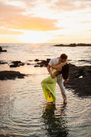 Couple sharing a romantic kiss at sunset, standing in shallow ocean water with rocky coastline and golden sky.