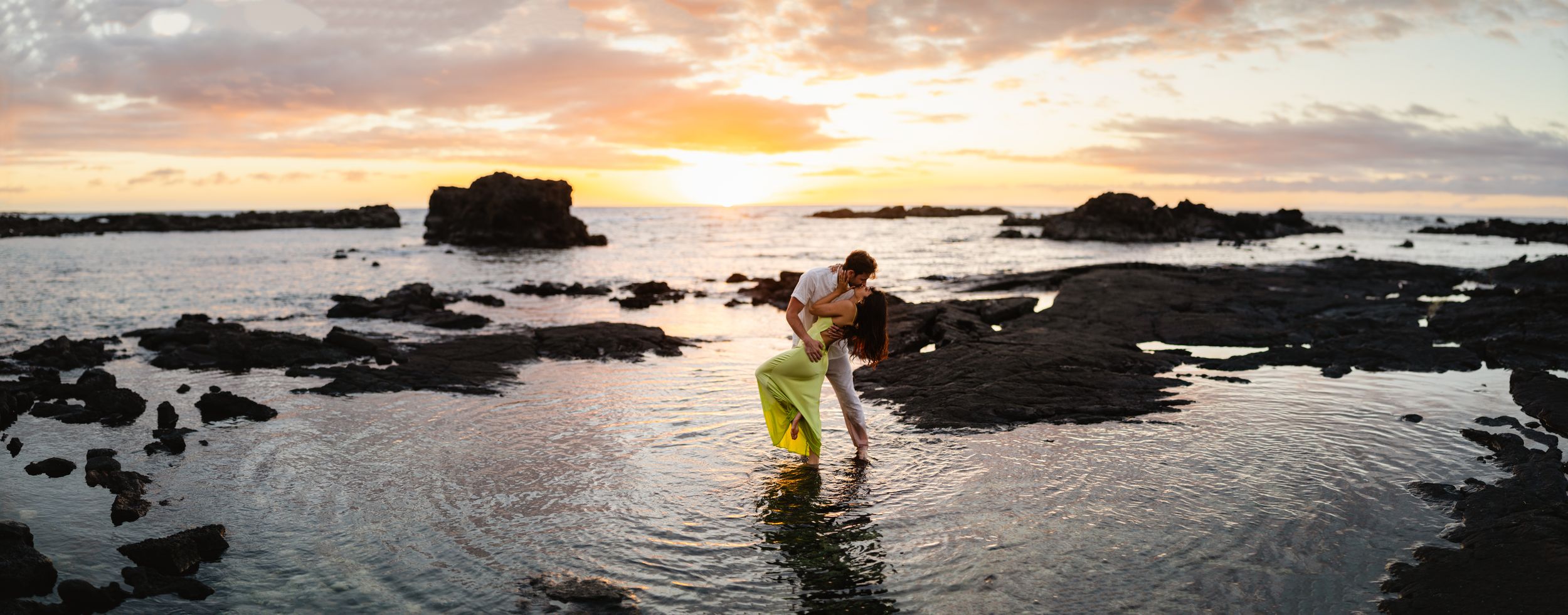 Couple embracing on rocky shoreline during golden sunset, waves reflecting warm orange hues in a romantic coastal scene.