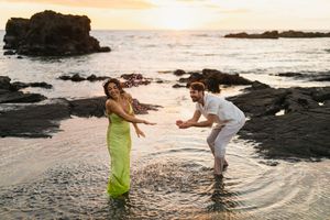 Couple laughing and splashing in ocean water at sunset near rocky shoreline, woman in green dress, man in white outfit.