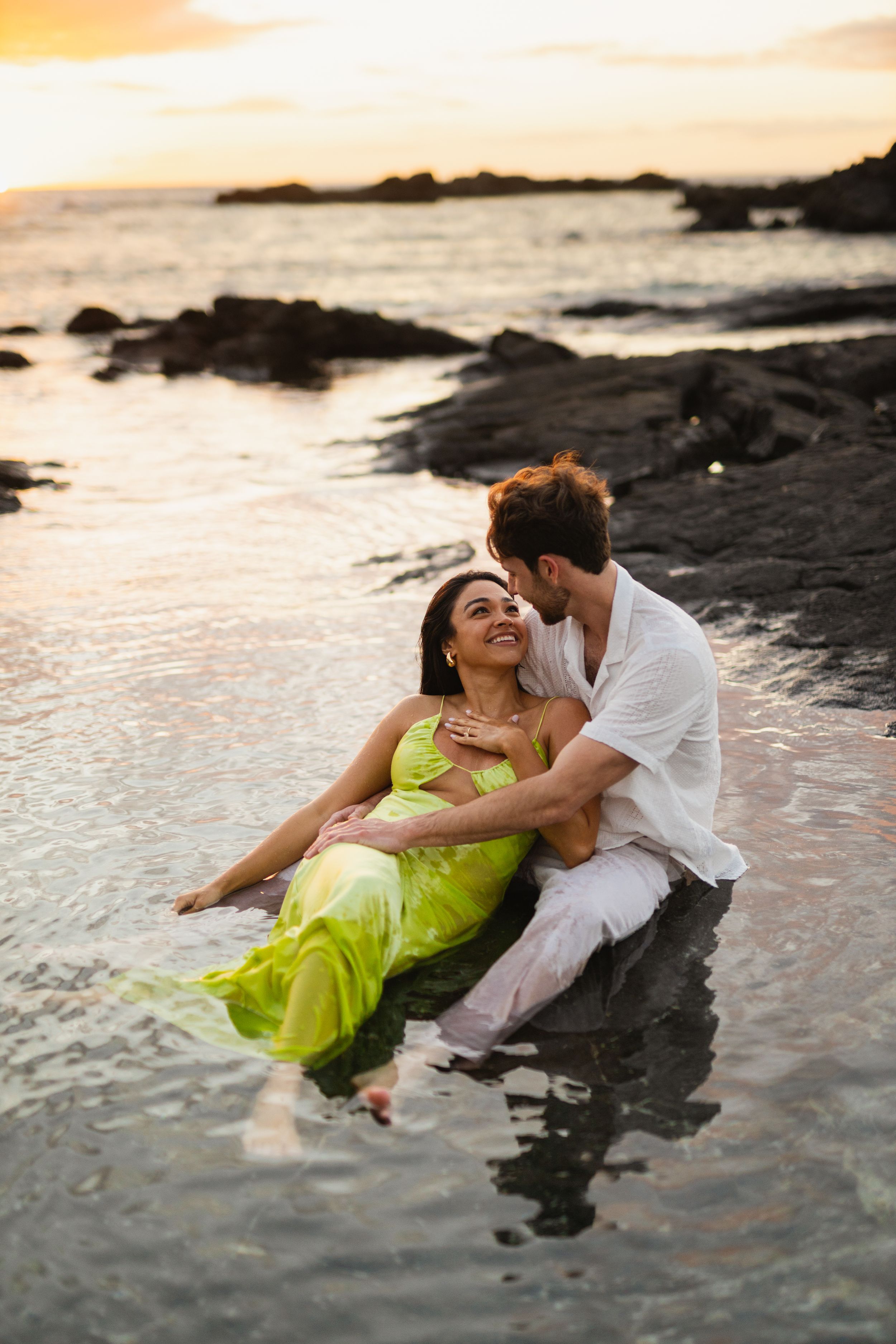 Romantic couple sitting in shallow ocean water at sunset, woman in green dress, man in white shirt embracing her.