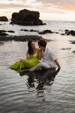 Romantic couple sitting in shallow ocean water at sunset, woman in green dress, man in white shirt, rocky coastline backdrop.