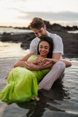 Romantic couple sitting in shallow ocean water at sunset, woman in green dress leaning back into mans embrace on rocky beach.