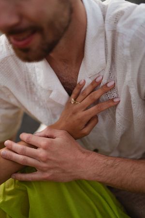 Close-up of a couple holding hands, woman wearing a gold ring, man in white linen shirt and green pants.