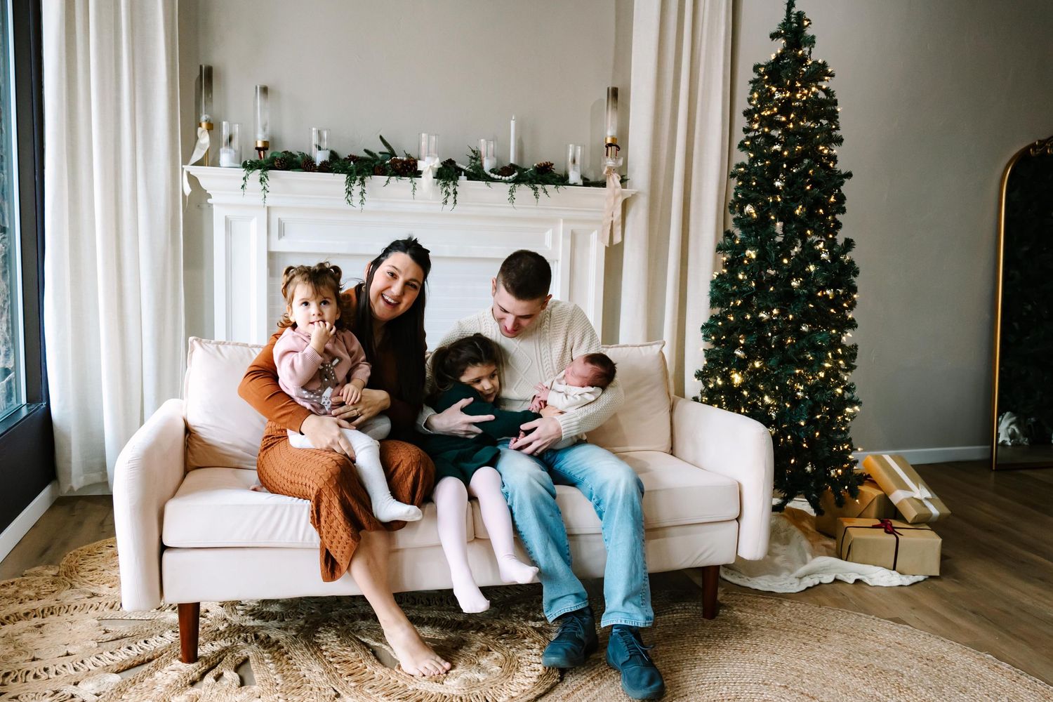 A family relaxes together on a white couch in front of a decorated Christmas tree and mantle with holiday decor.