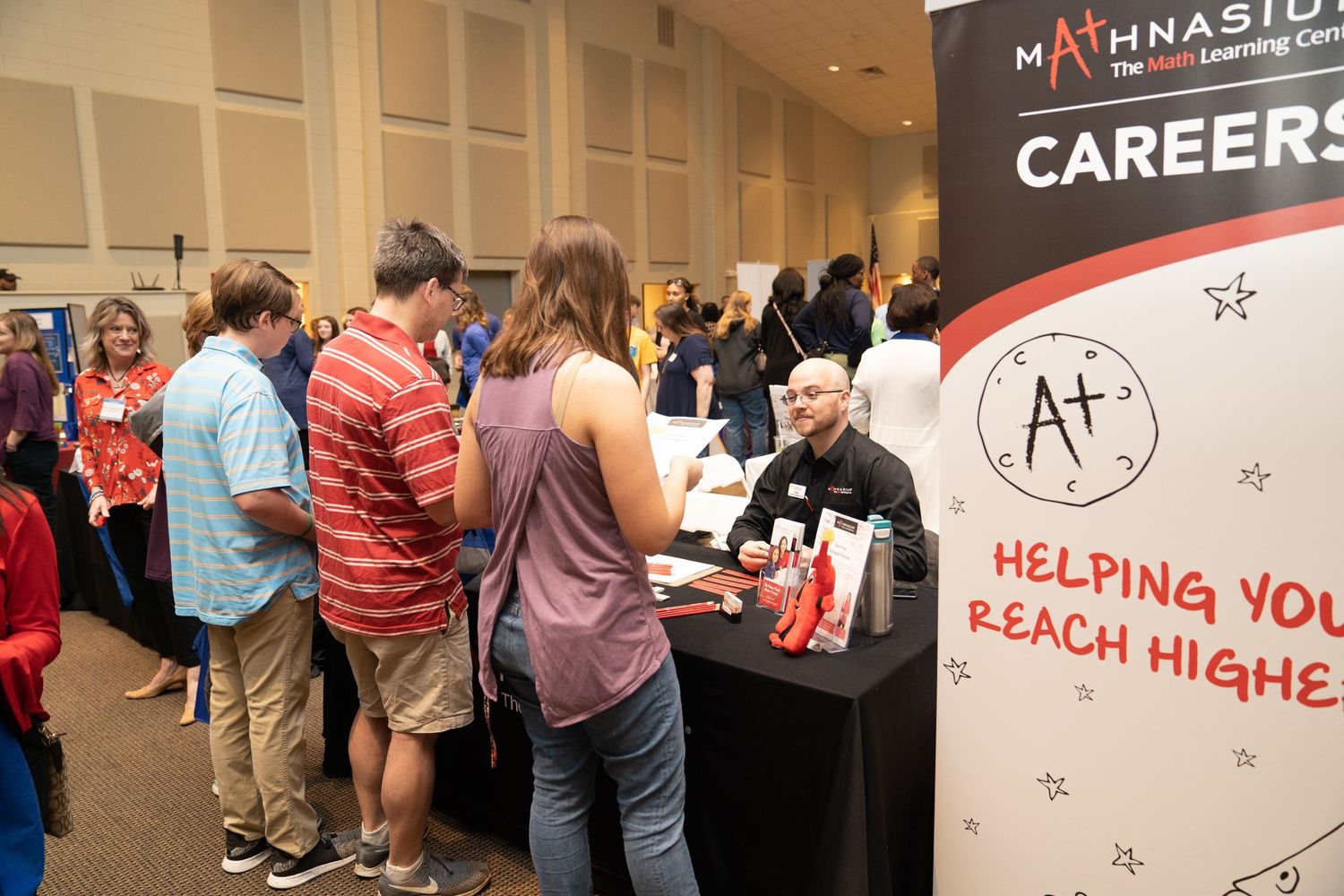 Students speak with Mathnasium rep at a career fair booth promoting tutoring jobs and education careers with “Helping You Reach Higher” bann