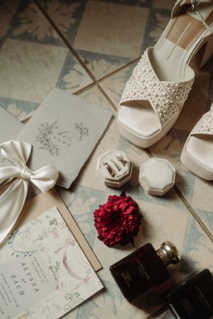 Wedding details flatlay with cream lace platform sandals, floral invitation with bow, red flower and perfume bottle.