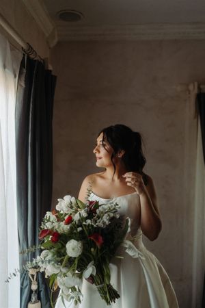 Elegant bridal scene by window with white gown and romantic red and white floral bouquet in natural light.