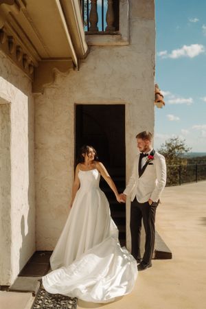 A couple in wedding attire stands at a stucco doorway with a flowing white wedding dress train on a sunny balcony.