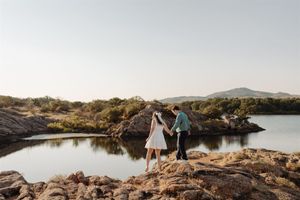 A couple stands together on rocky terrain overlooking a serene lake during golden hour.