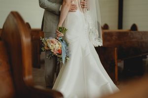 A bride in an elegant white wedding gown holds a colorful bouquet while standing in a church aisle.