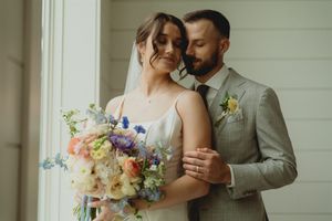 A romantic wedding portrait showing an embracing couple with a colorful spring bouquet by a window.