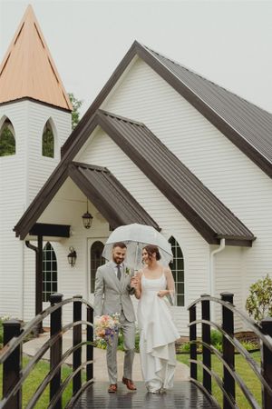 A white wooden chapel with a copper steeple and dark metal roof features a couple under an umbrella on a rainy day.