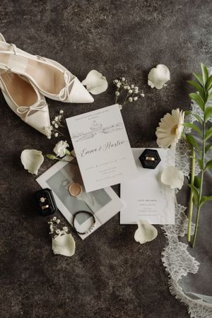 Wedding details flatlay with white shoes, invitation, flowers and rings on dark background.