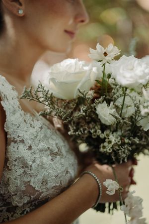 A delicate white bridal bouquet with roses and lace dress detail shown in closeup.