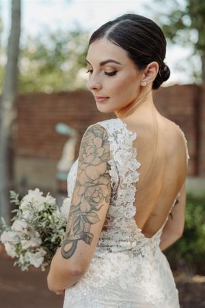 A bride in a backless lace wedding dress showcases her floral arm tattoo while holding a white bouquet.