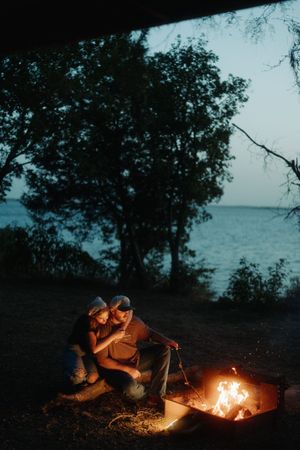 Couple sitting by a campfire at dusk near a lake with trees silhouetted against the darkening sky.
