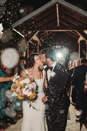 Couple sharing romantic kiss under covered venue with confetti falling during wedding celebration.
