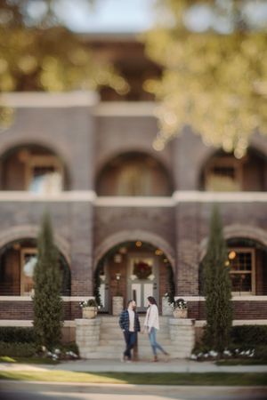 A couple stands in front of a brick building with arched windows and cypress trees framing the entrance.