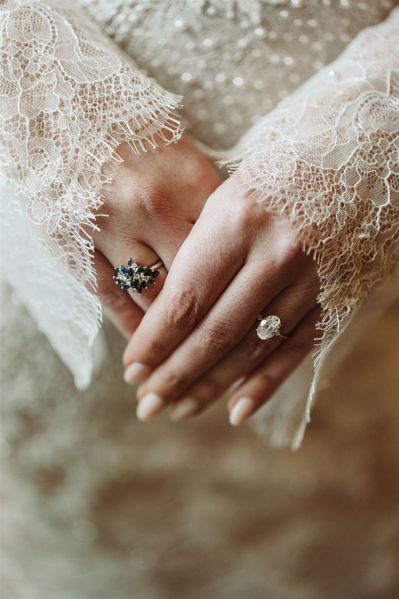 Two delicate rings on hands draped in white lace, featuring a blue gemstone and clear stone design.