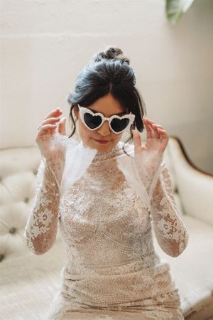 A bride wearing a lace long-sleeve wedding dress and heart-shaped sunglasses sits on a cream couch.