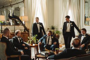 Groomsmen in tuxedos gather in elegant room with white curtains and vintage furniture for pre-wedding socializing.