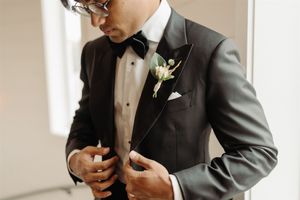 A figure in a black tuxedo and bow tie adjusts their buttons, wearing stylish glasses.