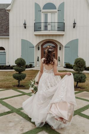 A bride in a flowing lace wedding dress stands in front of a white house with pale blue shutters and manicured topiaries.
