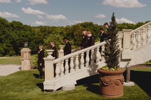 Two people stand on ornate stone staircase surrounded by landscaped gardens and decorative potted plants.