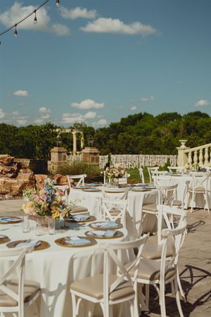Elegant outdoor wedding reception setup with white tablecloths, cross-back chairs and string lights under blue summer sky.