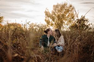 A couple shares an intimate moment while sitting in tall brown grass during golden hour at sunset.
