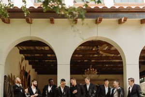 Row of groomsmen in black tuxedos stand under white Spanish-style arches at a wedding venue.