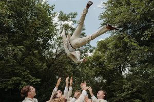 A group tosses a person wearing white clothing into the air in a dramatic celebratory moment surrounded by trees.