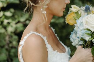 Close-up detail of lace wedding dress with blue and yellow floral bouquet against green garden background.