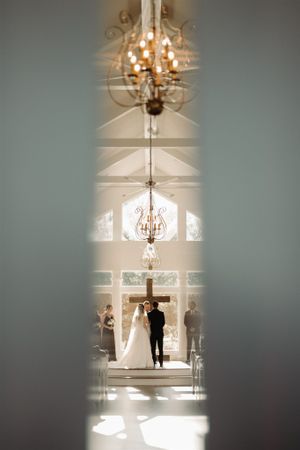 Wedding ceremony viewed through a doorway with crystal chandeliers illuminating the elegant space.