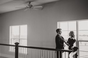 A couple shares an intimate black and white moment standing near large windows in an upstairs hallway with a ceiling fan.