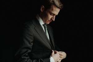 Professional in dark suit and green tie adjusts cuffs while standing in dramatic shadow lighting.