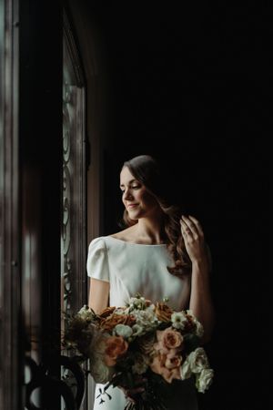 Elegant figure in white dress stands by vintage window holding romantic peach and cream floral bouquet.